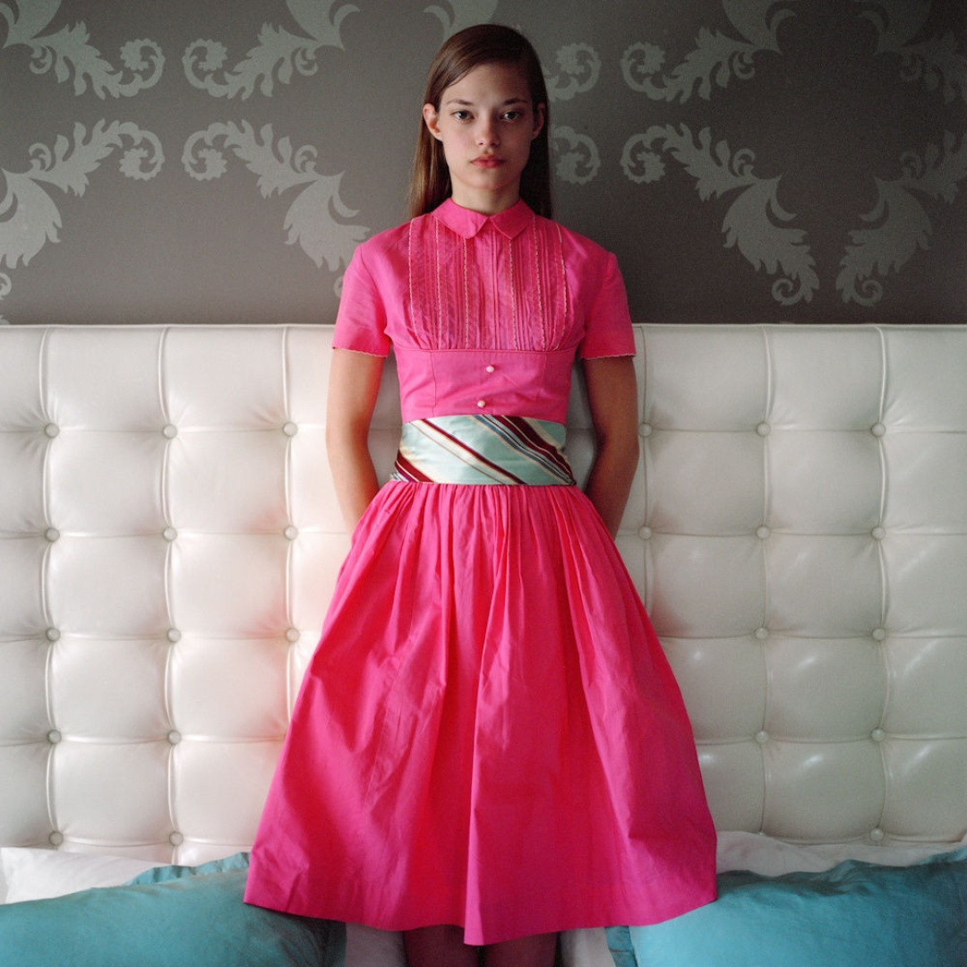 Model wearing a pink dress with a belt, standing in front of a white tufted headboard.